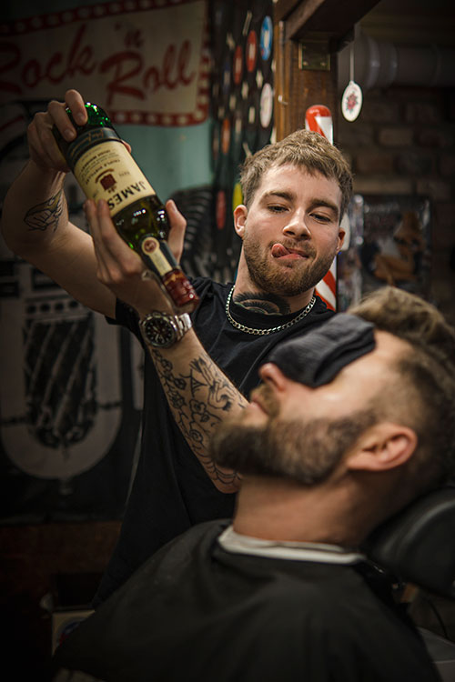Żartobliwy barber z butelką whiskey pozujący nad klientem z zasłoniętymi oczami, w barbershopie o rock'n'rollowej estetyce.