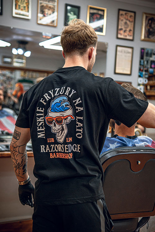 Barber w koszulce z napisem 'Męskie fryzury na lato' odwrócony tyłem do kamery, pracujący w tle pełnym barbershopowych memorabiliów.