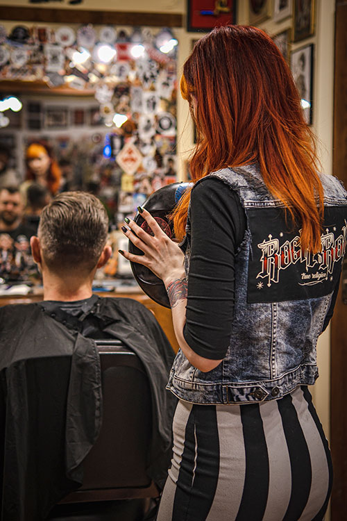 Rudowłosa kobieta w stylowej kamizelce obserwująca efekt swojej pracy w barbershopie, trzymając w dłoniach okrągłe lustro.