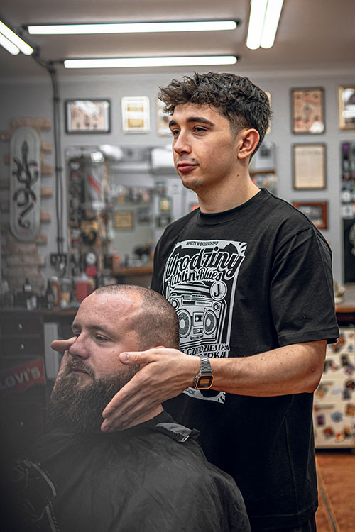 Uważny młody barber dokładnie stylizujący brodę klienta, z odbiciem profesjonalnej atmosfery barbershopu w lustrze.