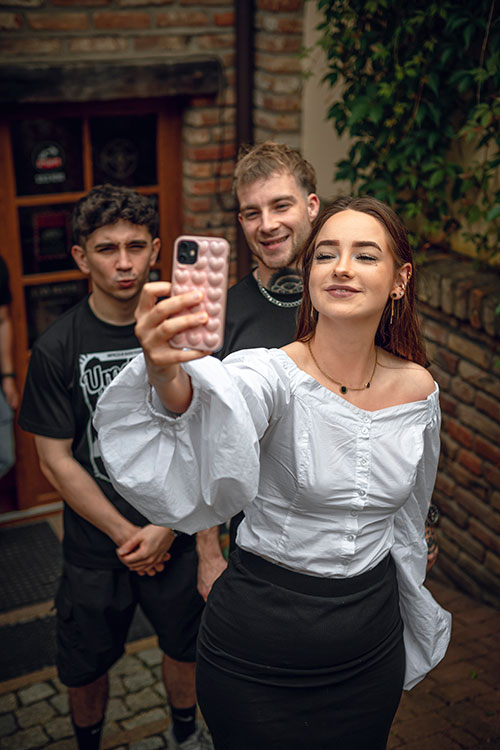Uśmiechnięta młoda kobieta robi selfie z dwoma mężczyznami w tle, stojąc przed wejściem do barbershopu, promieniująca radością.