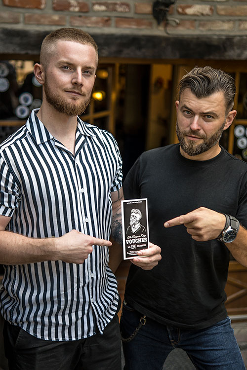 Dwóch przystojnych mężczyzn prezentujących voucher na usługi barberskie, z uśmiechem wskazują na logo barbershopu, promując lokalne usługi.