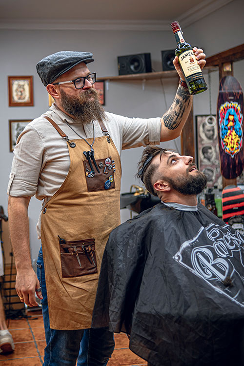Codzienność w salonie - Barber z brodą i okularami podnoszący butelkę whiskey nad głową klienta leżącego w fotelu, w żartobliwym i niekonwencjonalnym geście.