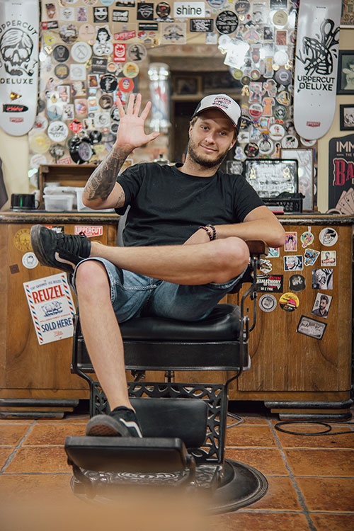 Przyjacielski barber w czapce machający do kamery, siedzący na klasycznym fotelu barberskim, otoczony żywą kolekcją naklejek i pamiątek barberskich na ścianie