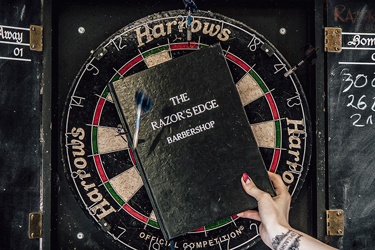 Tarcza do darta z trafionym środkiem obok czarnej książki z napisem 'The Razor's Edge Barbershop', sugerująca zabawną i konkurencyjną atmosferę.