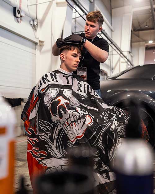 Strzyżenie w hali Moto Session – barber w akcji, peleryna z czaszką robi robotę.