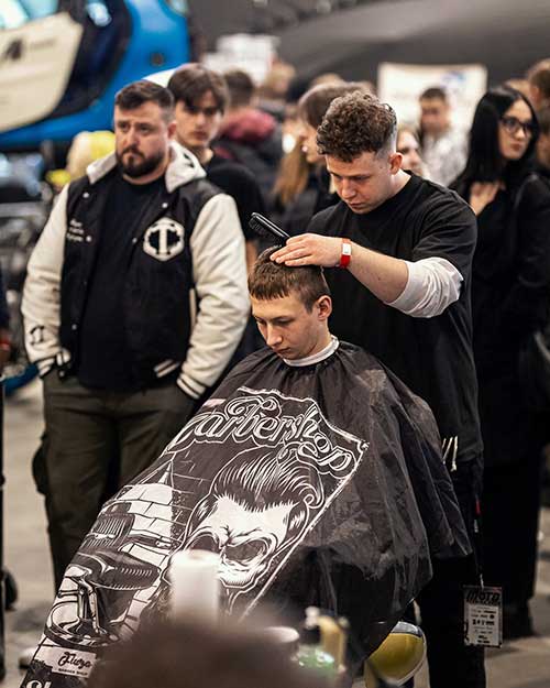 Młody gość na fotelu, barber skupiony, a za nimi tłum i motoryzacyjny zgiełk.