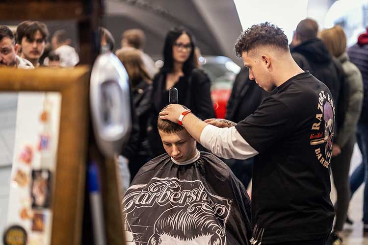Młody klient na fotelu, fryzjer z Razor’s Edge skupiony przy cięciu, a za nimi zainteresowani goście.