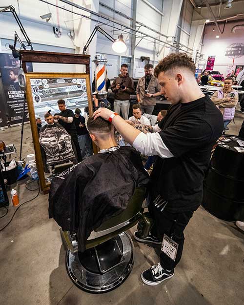 Strzyżenie przy naszym stanowisku – fryzjer w pełnym skupieniu, wokół aparat, banery i widzowie.