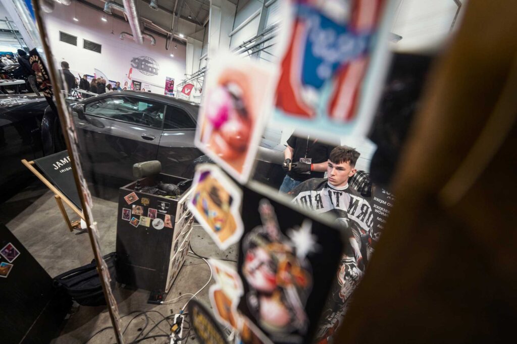 Nieoczywisty kadr przez naklejki na lustrze – w tle klient i barber w skupieniu.