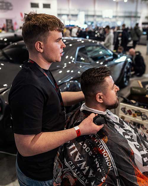 Barber trzyma maszynkę przy szyi klienta – z tyłu luksusowe auta, mocny kontrast stylów.