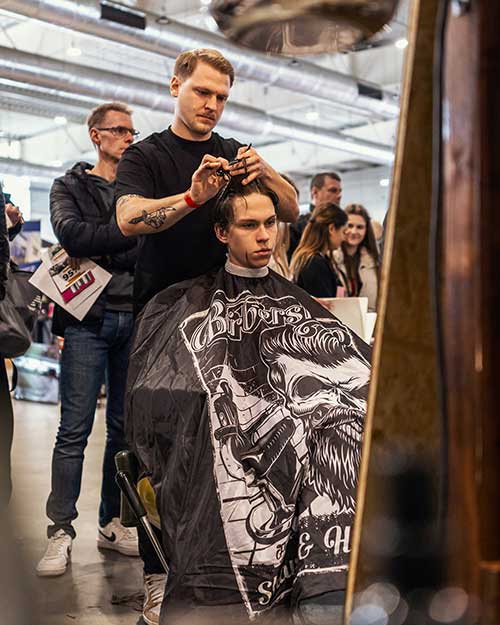 Barber tnie, klient w pelerynie z czaszką, a wokół publika – energia na stanowisku.