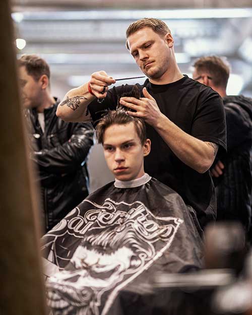 Młody klient z poważnym spojrzeniem, barber kończy fryzurę w zatłoczonej hali.