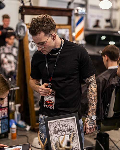 Tatuaże, identyfikator i pełne skupienie – barber ogarnia coś przy stole.